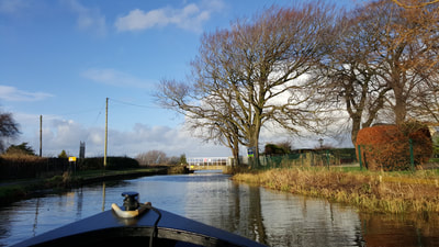 Bow of Narrowboat in foreground looking out onto one of the Lancaster canal swing bridge, with trees and canal scenery. Image links to things to do on lancaster canal