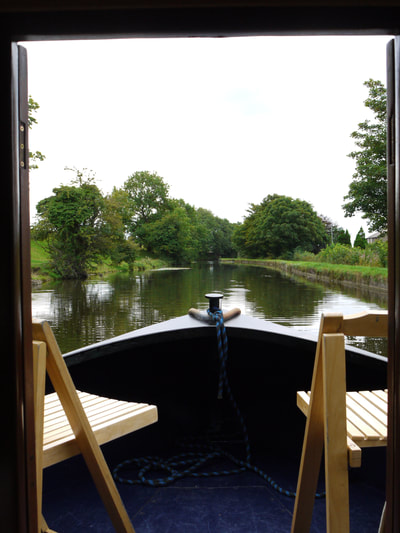The bow of a hire narrowboat looking out onto the Lancaster canal with green trees and canal towpath in the background